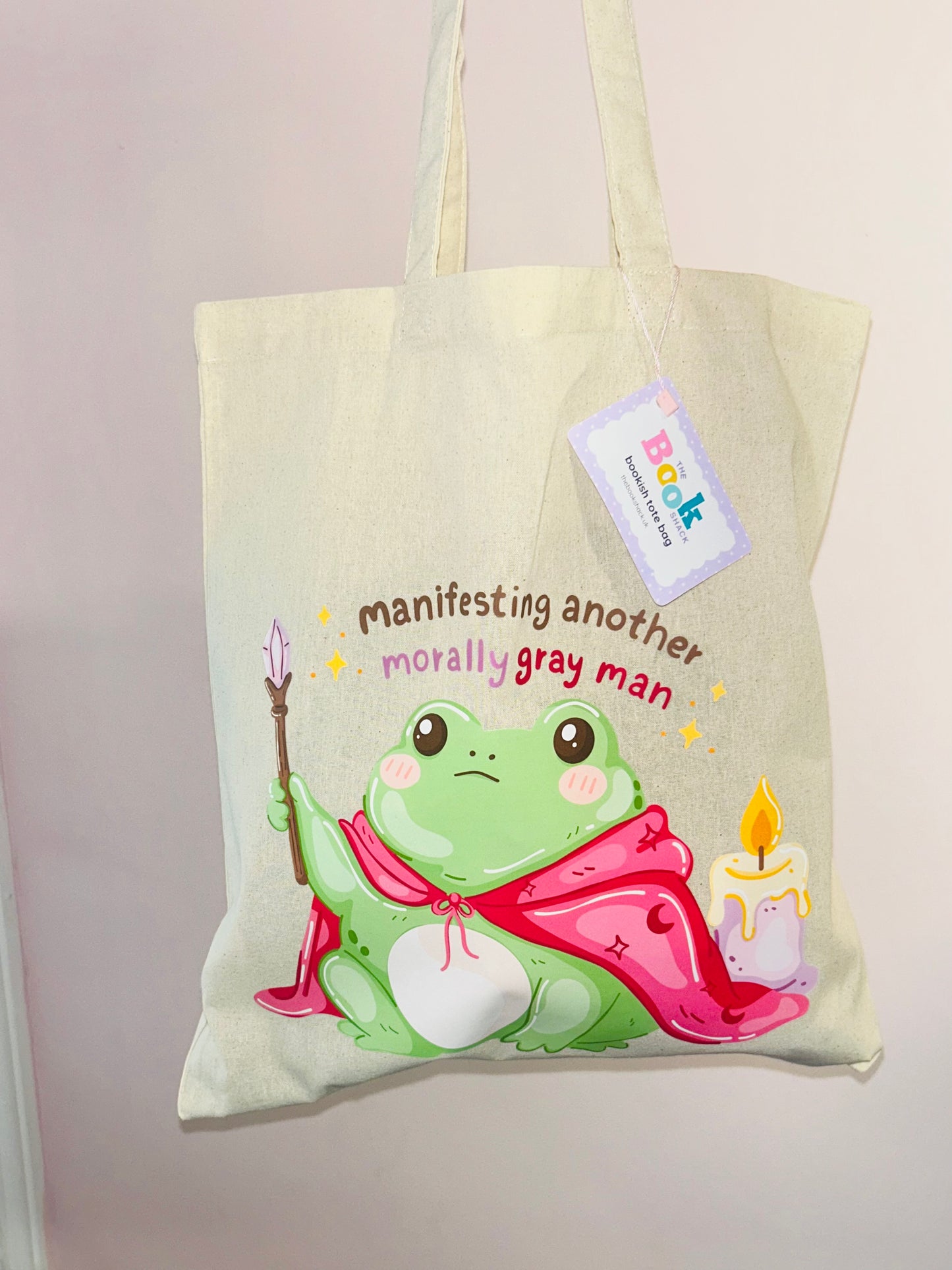 Cream canvas tote bag with seated cartoon frog, candles, and dangling price tag