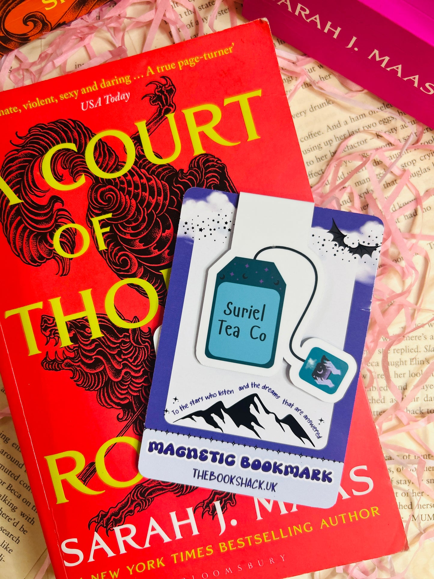 Blue tea bag-shaped magnetic bookmark labelled “Suriel Tea Co” clipped to an open fantasy novel, styled with pink shredded paper.