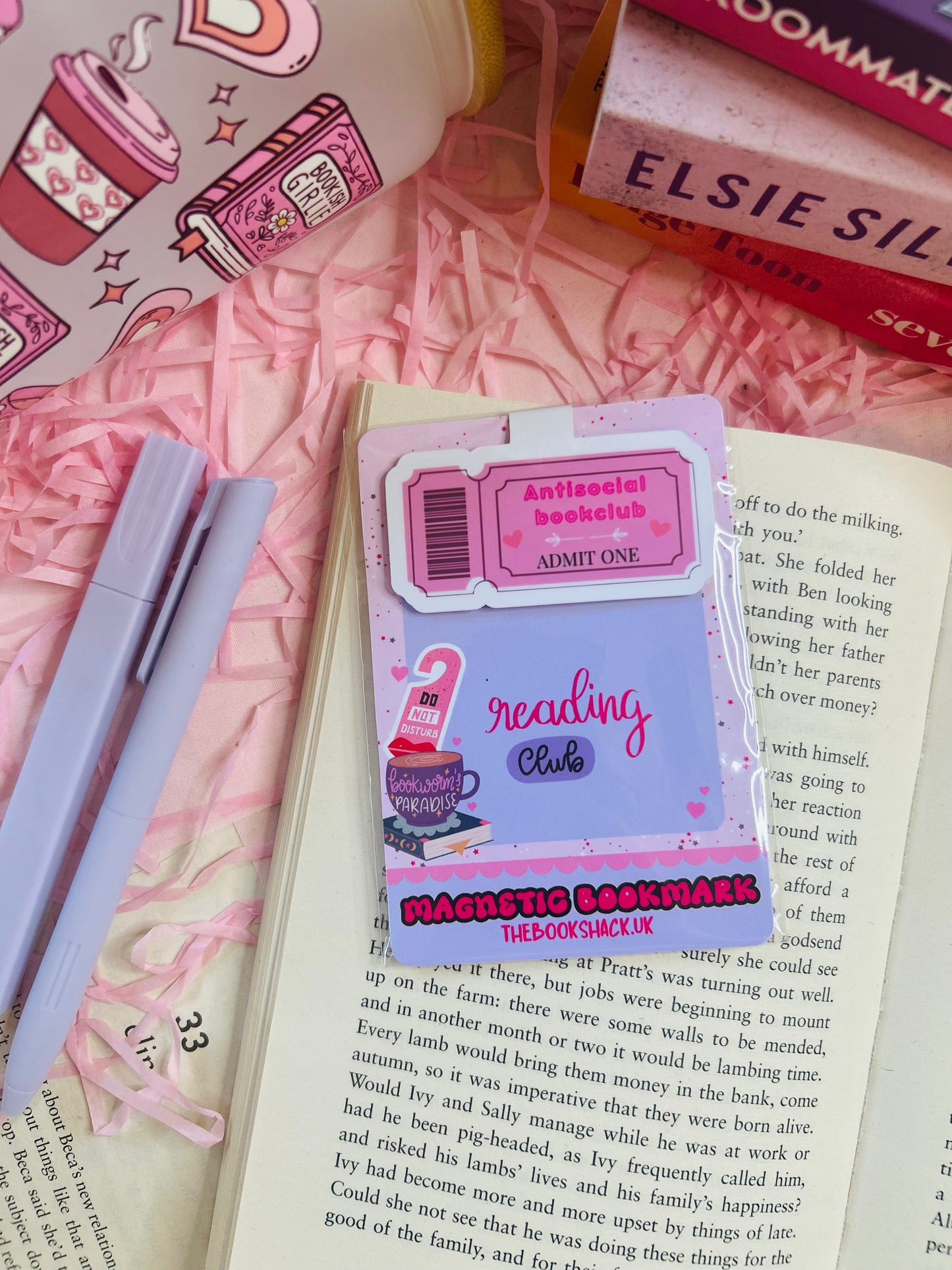 Pink magnetic bookmark with ‘Antisocial Book Club’ design on top of an open book, with pastel pens, shredded pink filler, and books in the background.
