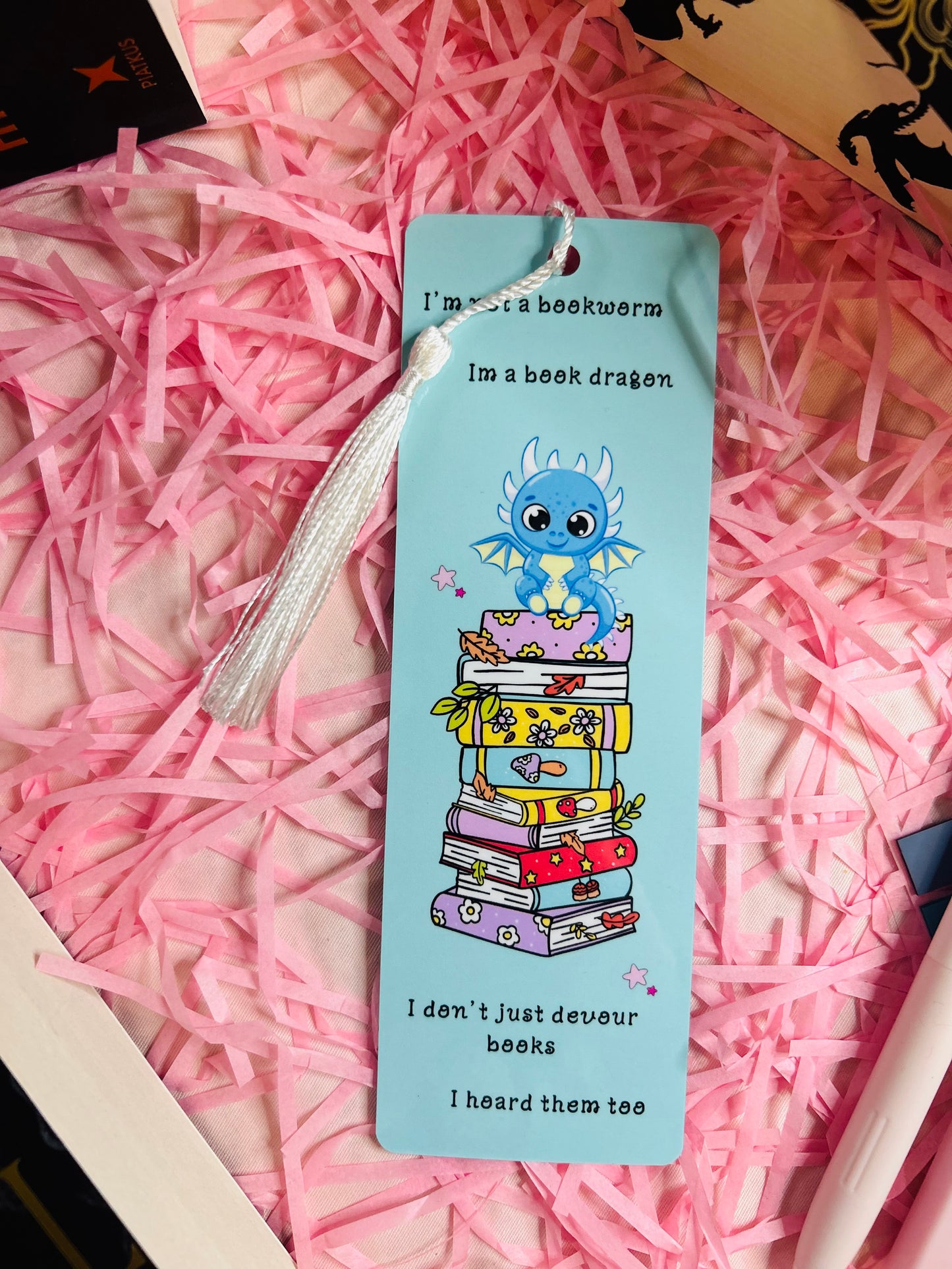 This updated bookmark now includes a delicate white tassel, adding a touch of elegance to the charming design. The whimsical dragon sitting atop a vibrant stack of books remains the focal point, paired with the playful quote: “I’m not a bookworm, I’m a book dragon. I don’t just devour books, I hoard them too.” The white tassel complements the light blue background, enhancing its appeal while maintaining functionality for avid readers