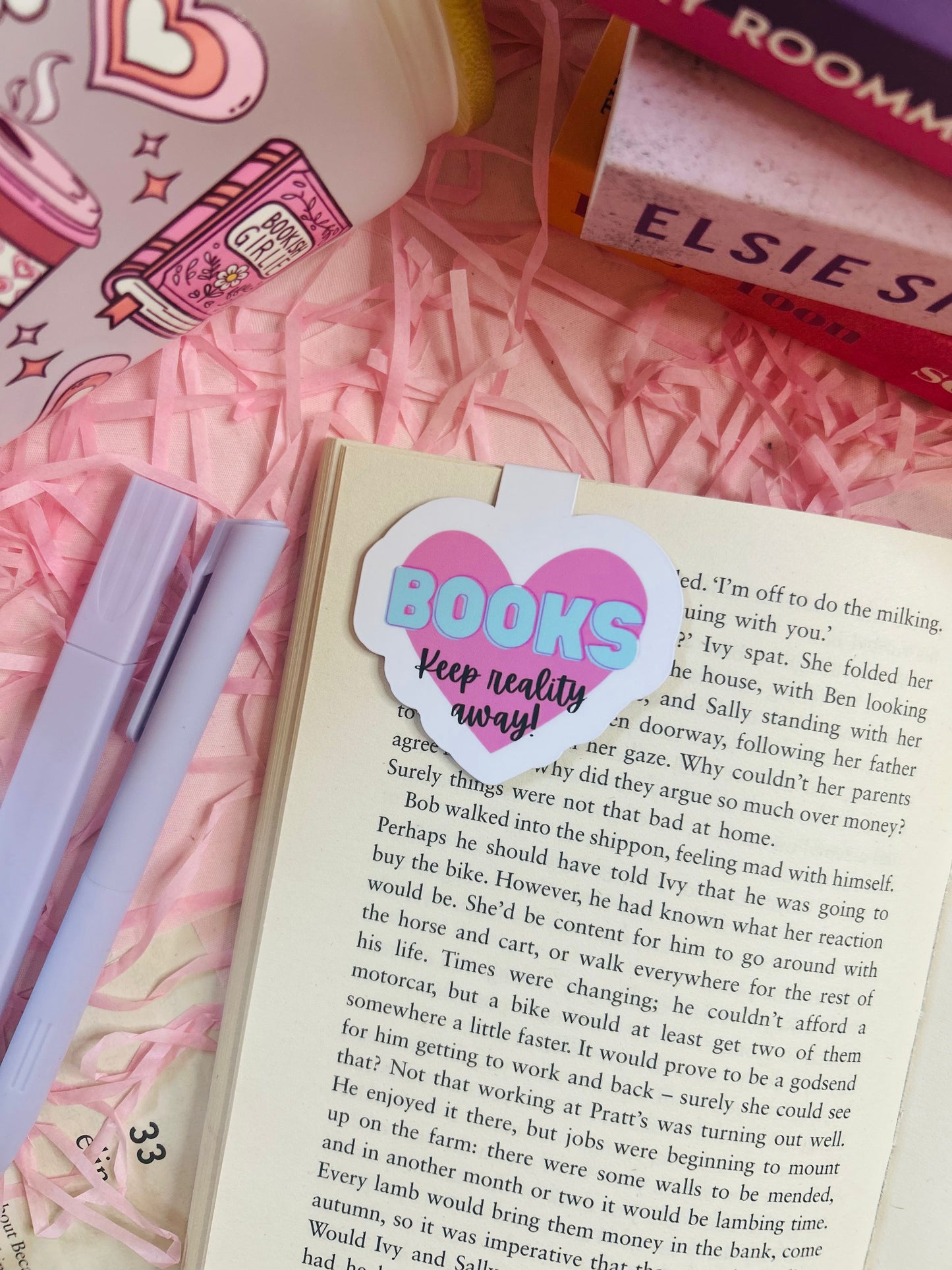 Heart-shaped pink magnetic bookmark with the phrase ‘Keep Reality Away’ on an open book surrounded by pink bookish accessories.