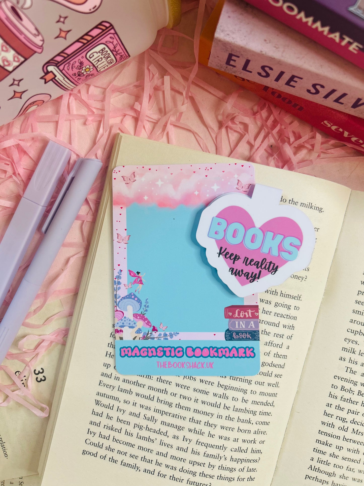 Heart-shaped pink magnetic bookmark with the phrase ‘Keep Reality Away’ on an open book surrounded by pink bookish accessories.