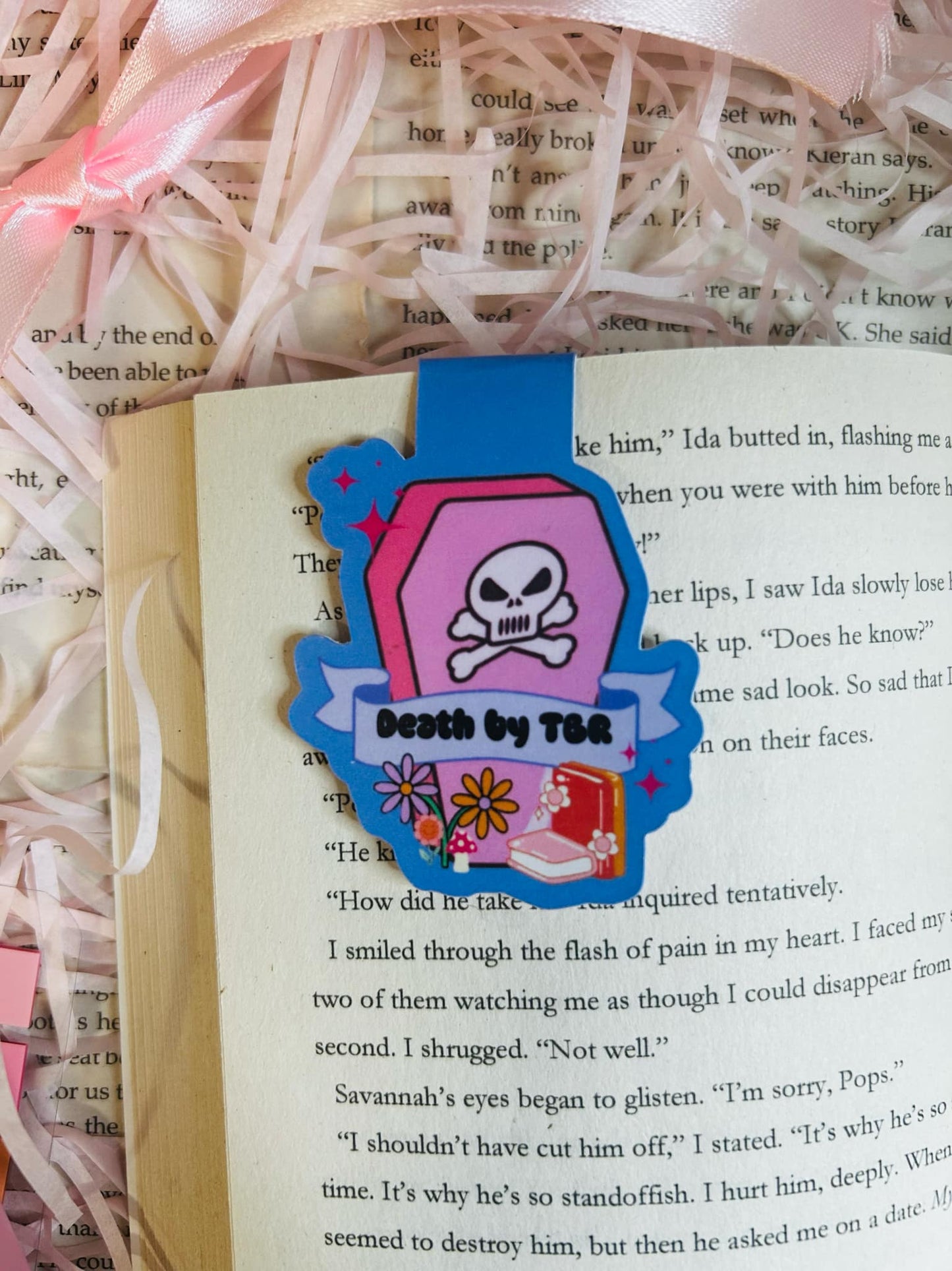 colourful 'Death by TBR' magnetic bookmark resting on an open book. The bookmark features a vibrant pink coffin design with a skull and crossbones, flowers, and a banner
