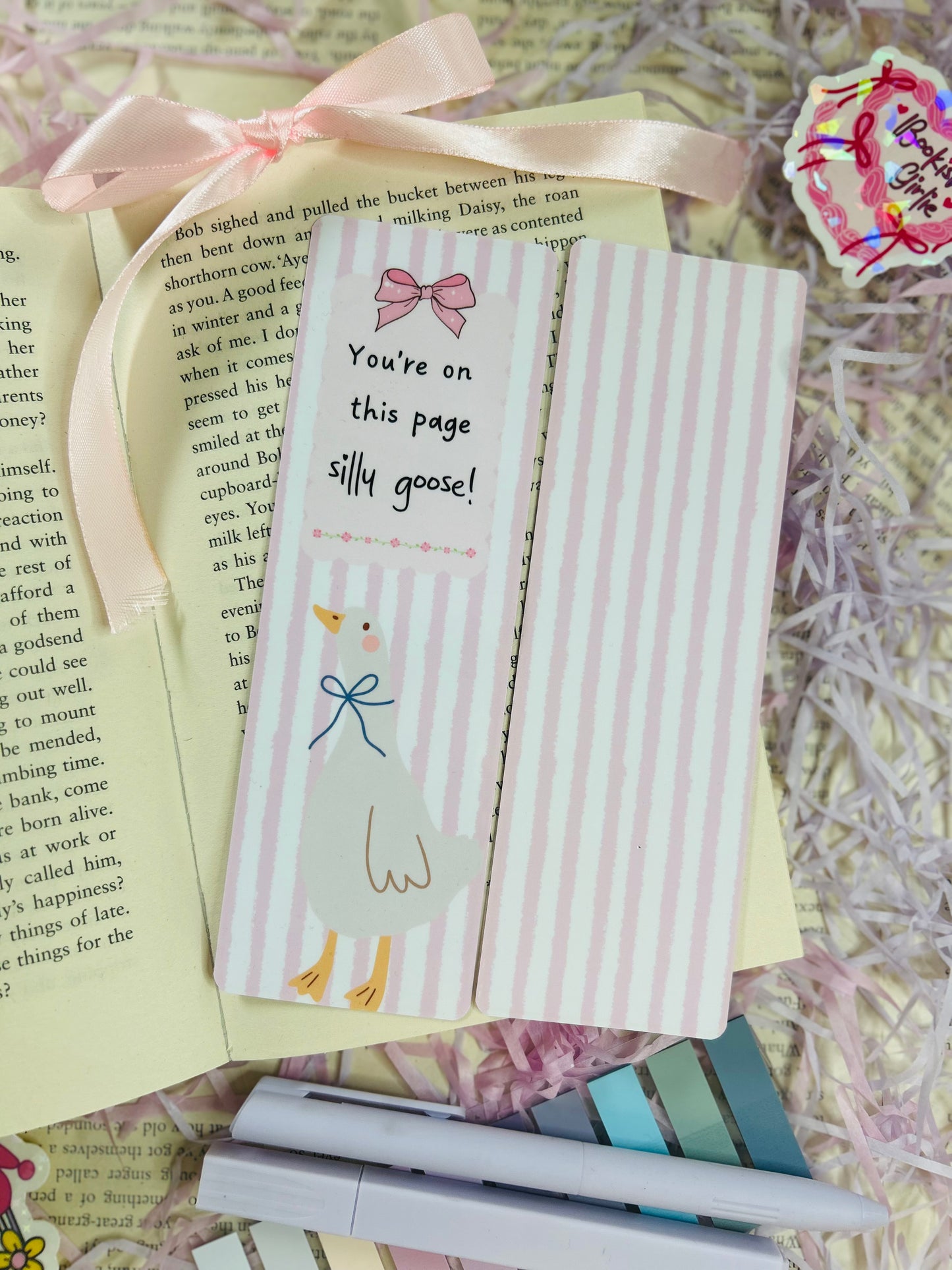 Set of three pastel bookmarks with silly goose and bow designs, placed on an open book with soft pink ribbon, shredded filler, and pens.