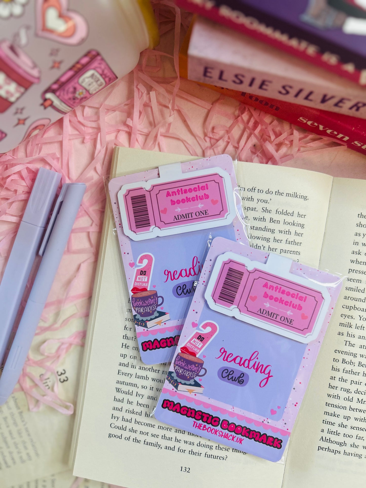 Pink magnetic bookmark with ‘Antisocial Book Club’ design on top of an open book, with pastel pens, shredded pink filler, and books in the background.
