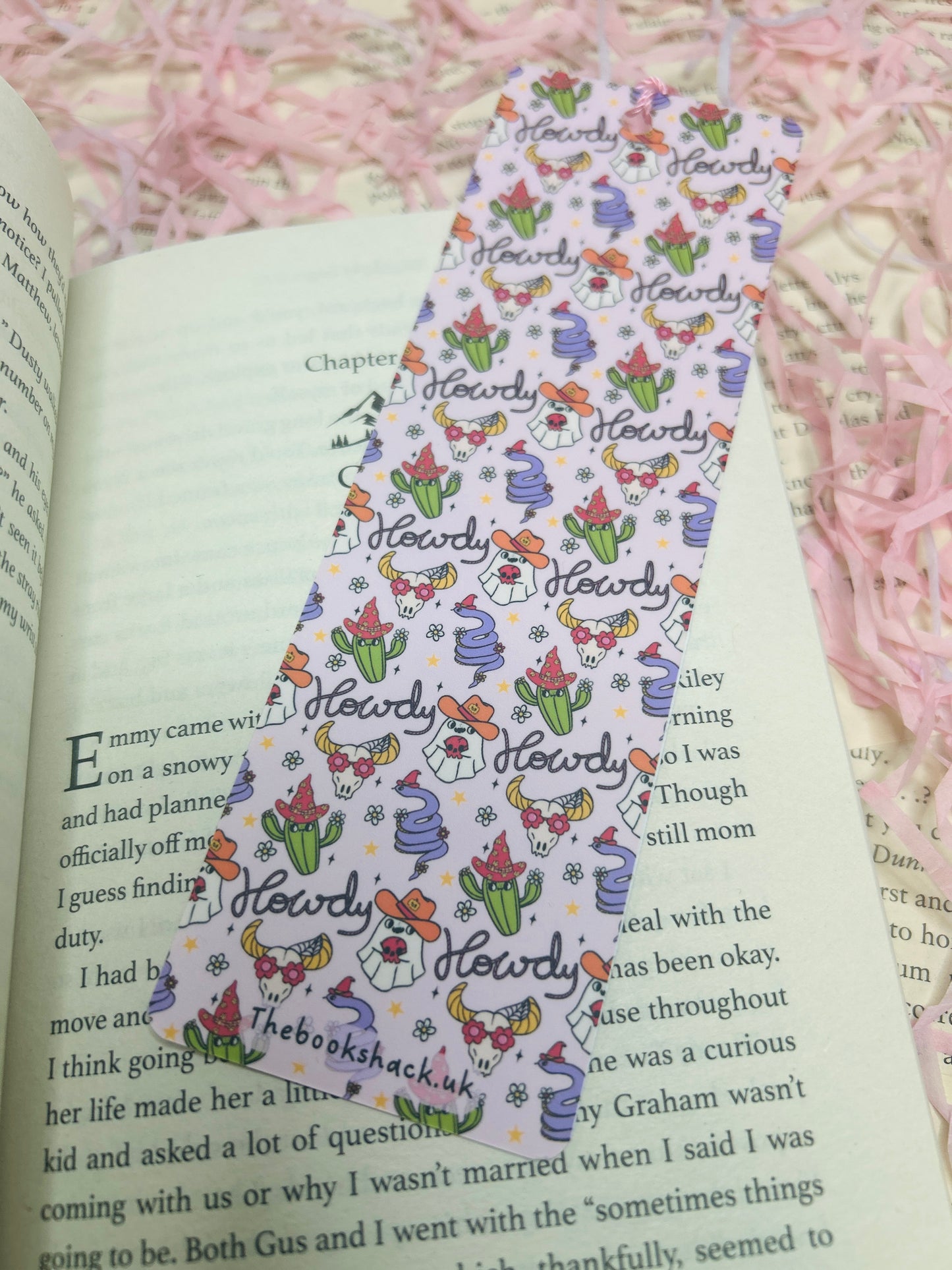 Double-sided western bookmark with repeating “Howdy” design featuring cowboy hats, boots, flowers, cacti, and cow-print accents, finished with a pink tassel.