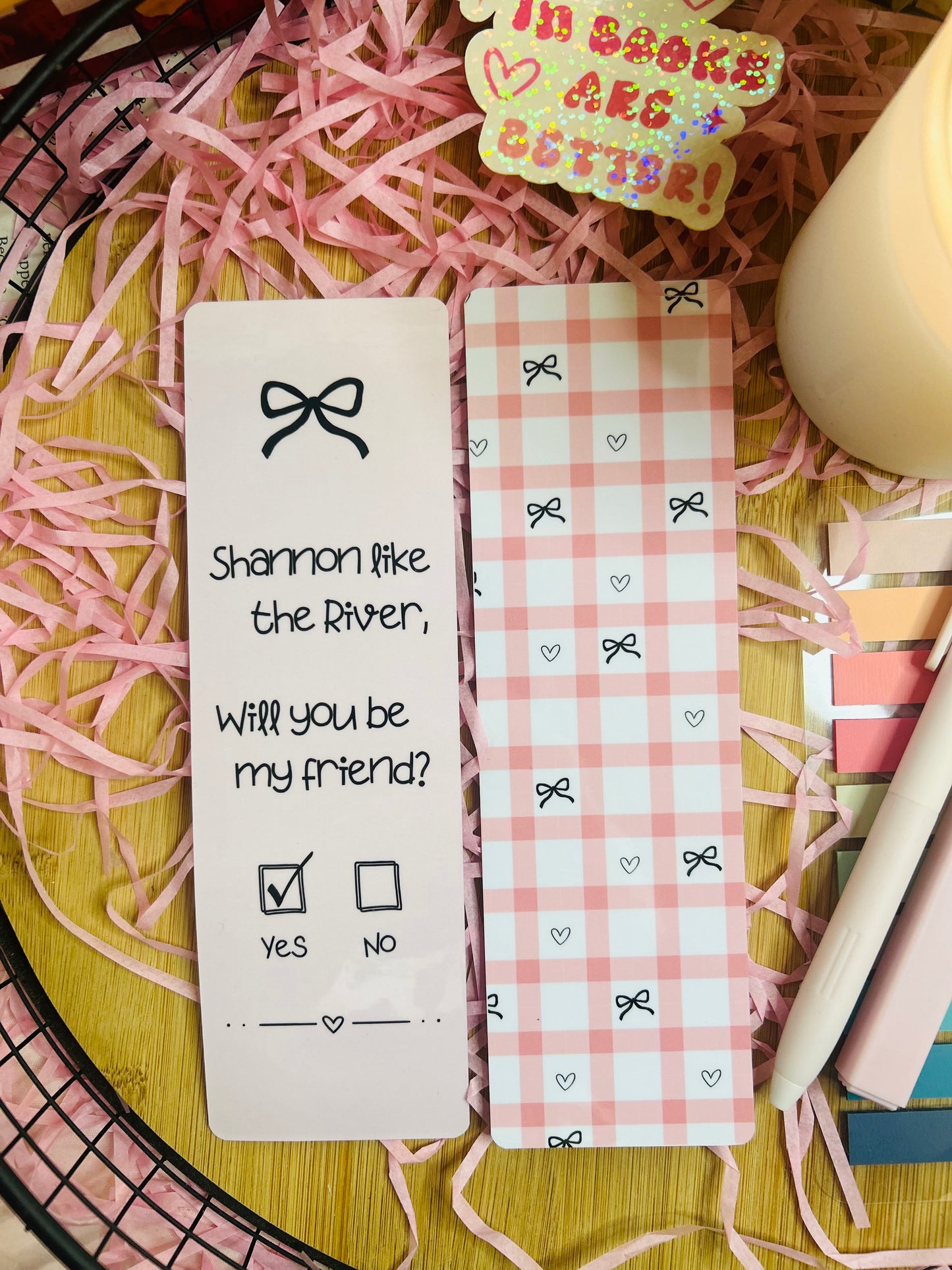 Two pink gingham bookmarks inspired by Binding 13, featuring a friendship note design with the phrase “Shannon like the River, Will you be my friend?” on one and a pink bow pattern on the other, styled with soft bookish decor.