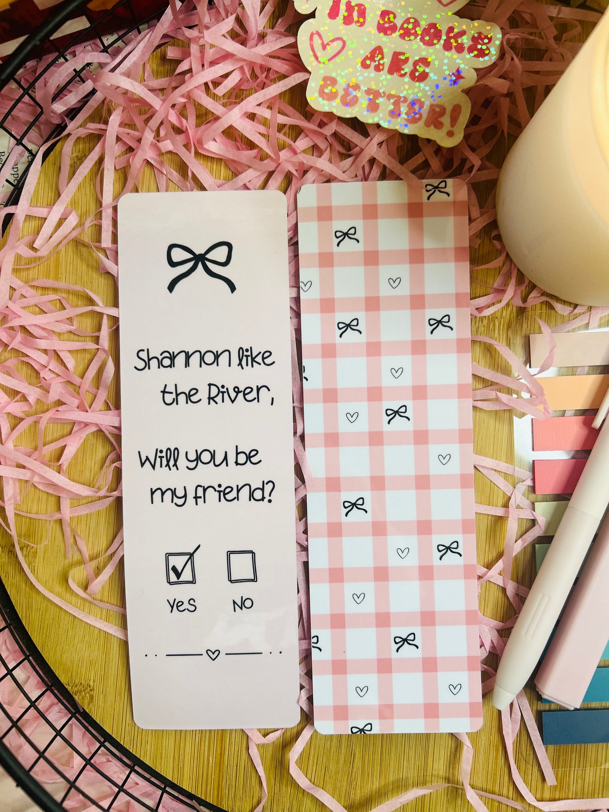 Two pink gingham bookmarks inspired by Binding 13, featuring a friendship note design with the phrase “Shannon like the River, Will you be my friend?” on one and a pink bow pattern on the other, styled with soft bookish decor.