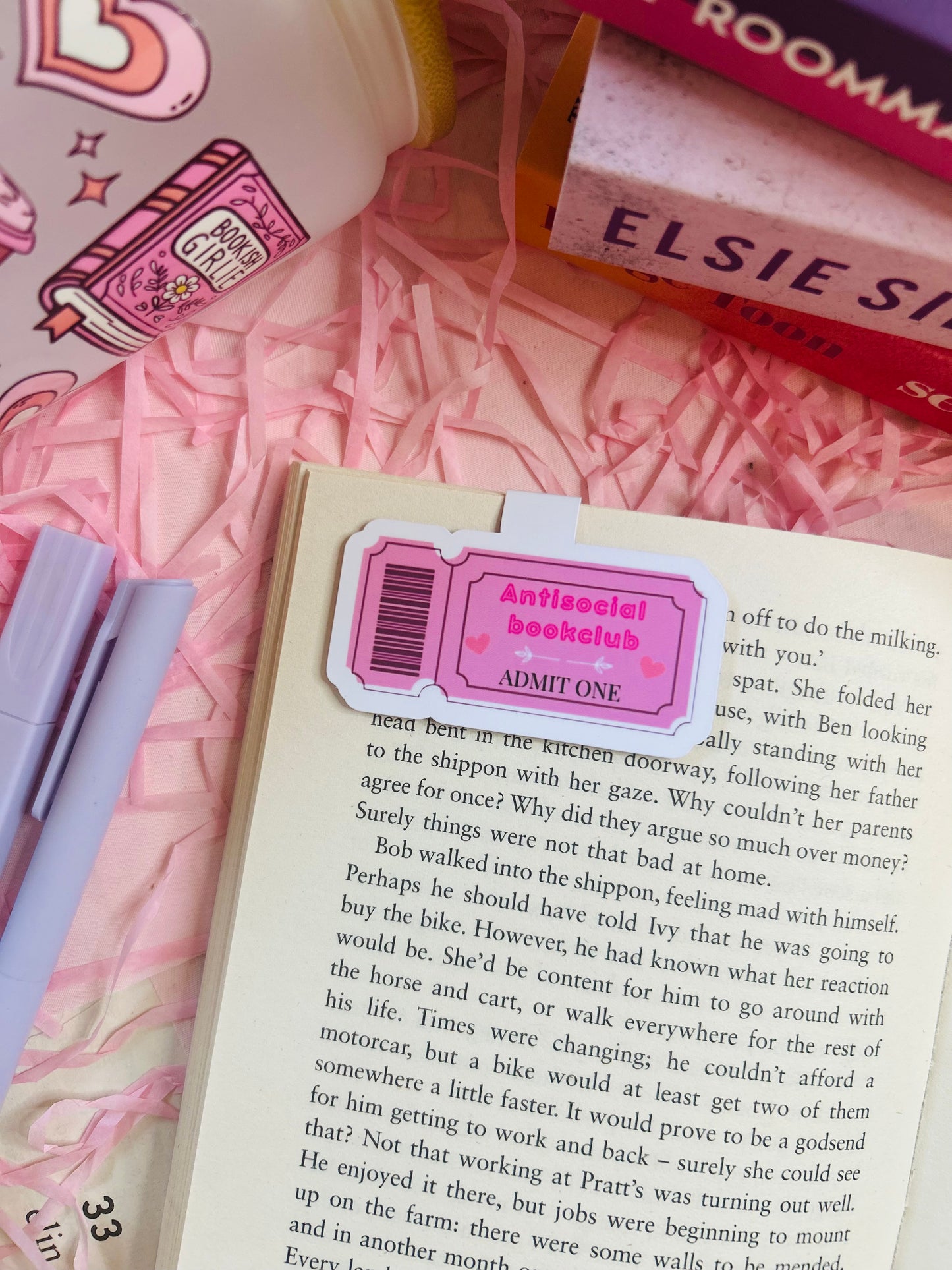 Pink magnetic bookmark with ‘Antisocial Book Club’ design on top of an open book, with pastel pens, shredded pink filler, and books in the background.