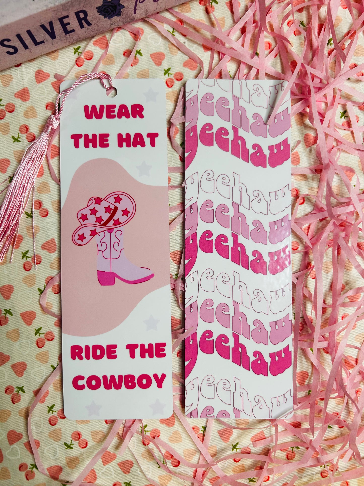 Western-themed bookmarks featuring a pink design with cowboy boots, hats, and bold text reading 'Wear the Hat, Ride the Cowboy' and a matching 'Yeehaw' pattern. Laminated bookmarks with tassels styled on a decorative pink and cherry background.