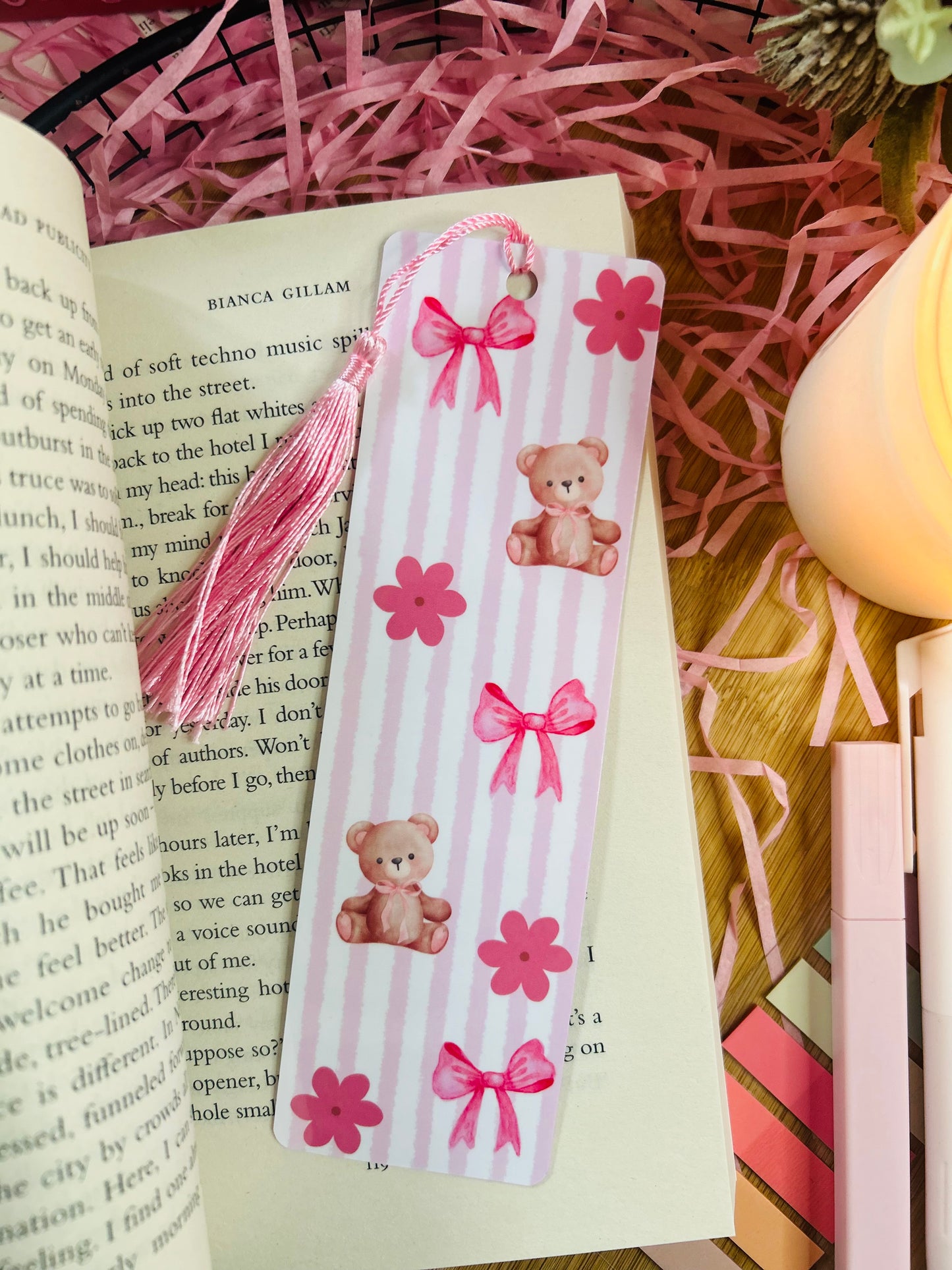 A pink teddy bear-themed bookmark with soft vertical stripes, delicate bows, and floral details, featuring a matching pink tassel.