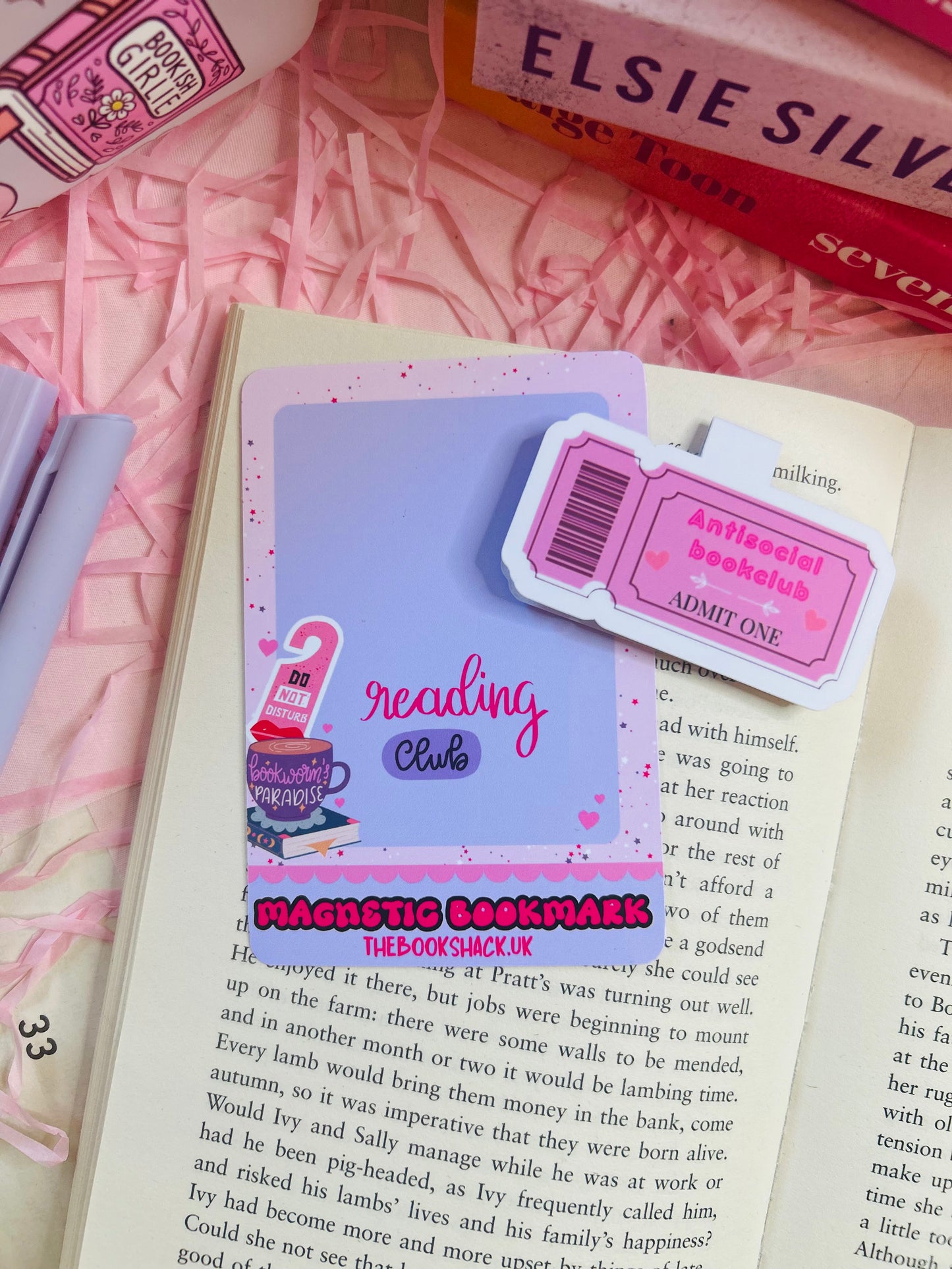 Pink magnetic bookmark with ‘Antisocial Book Club’ design on top of an open book, with pastel pens, shredded pink filler, and books in the background.