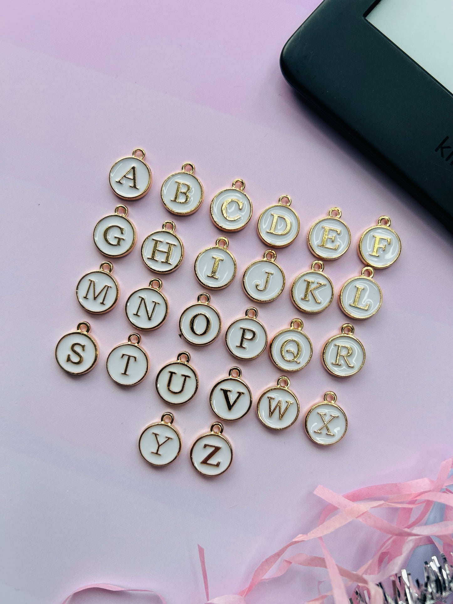 Alphabet letter charms on a pink surface with a phone in the background