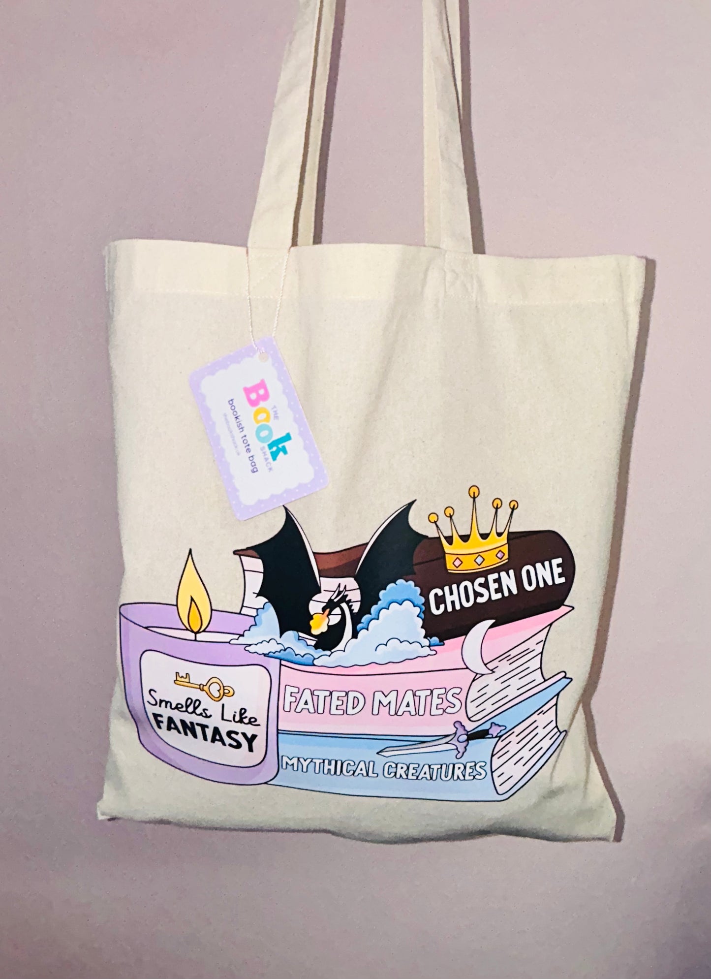 Cream tote bag with fantasy book stack graphic and hanging tag on handle.
