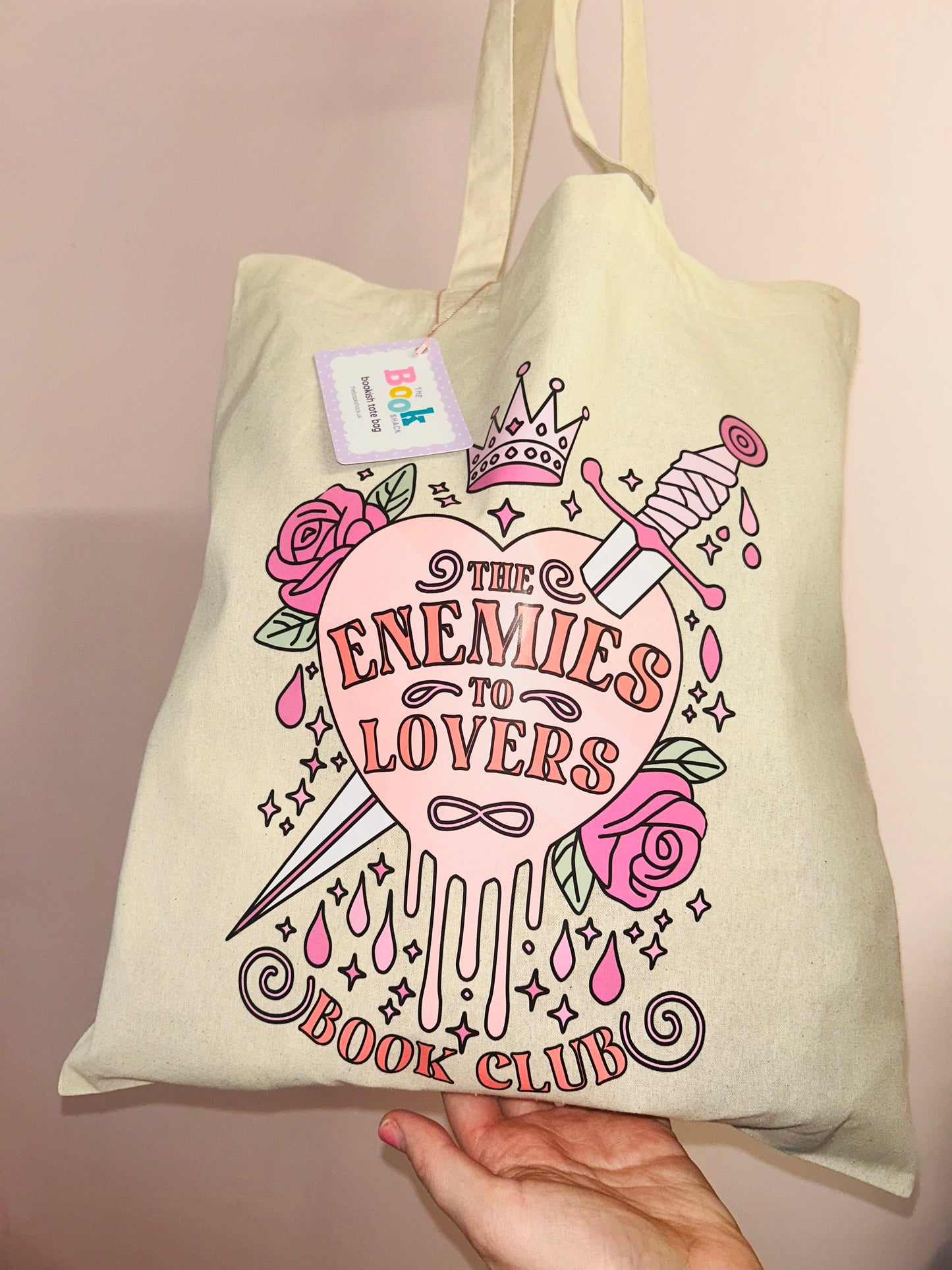 Cream cotton tote bag held in hand, pink “Enemies to Lovers” graphic with crown