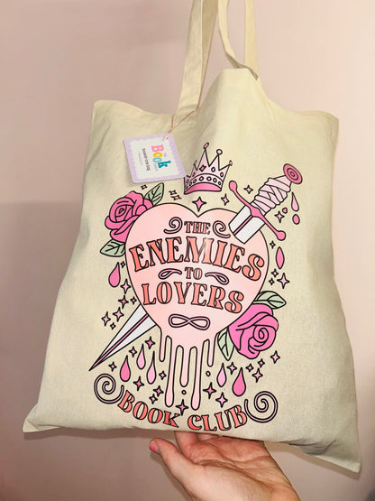 Cream cotton tote bag held in hand, pink “Enemies to Lovers” graphic with crown