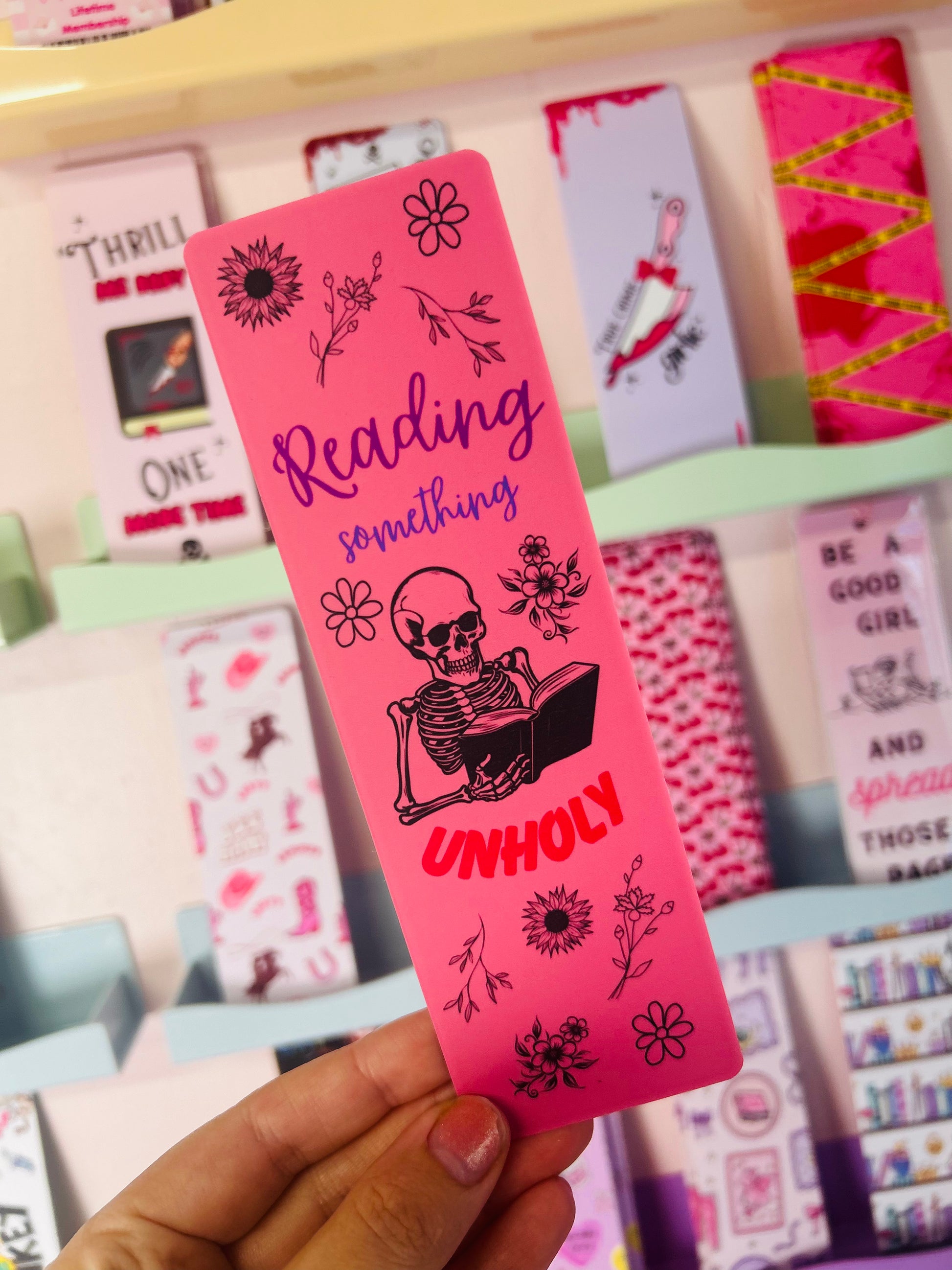 Pink bookmark held up, "Reading something" and "UNHOLY" with black skeleton and flowers