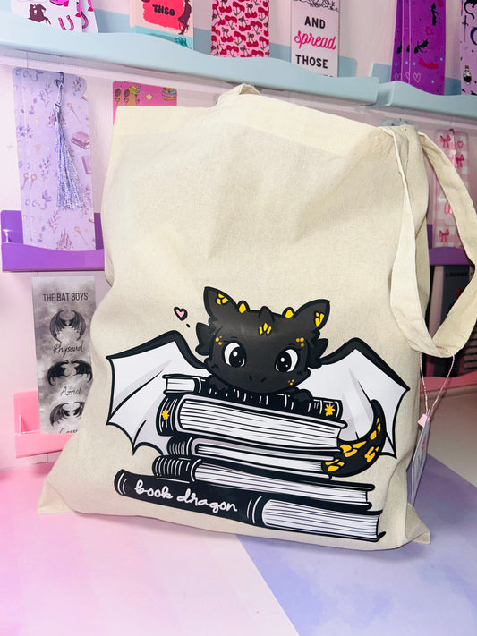 Cream cotton tote bag with black book-dragon graphic on front, top handles.