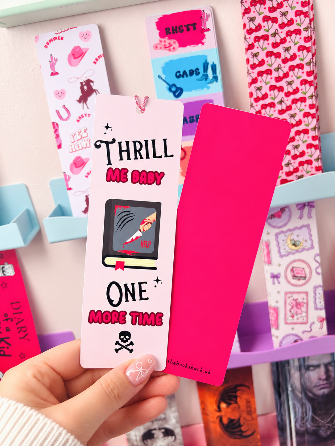 Pink tassel bookmark reading 'THRILL ME BABY' and 'ONE MORE TIME', held with another bookmark.