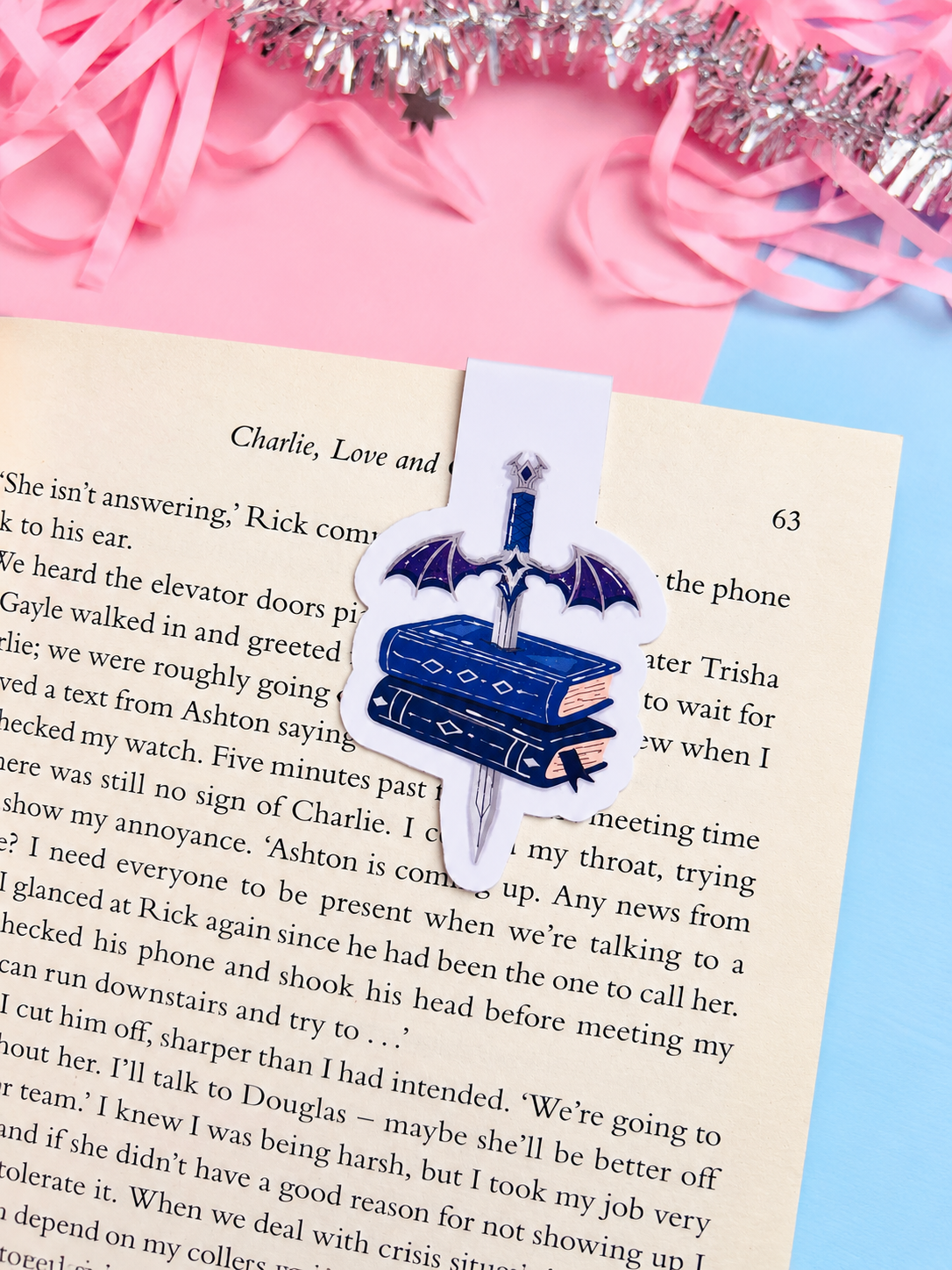 Magnetic bookmark shaped like a blue dagger over stacked books, clipped to an open page