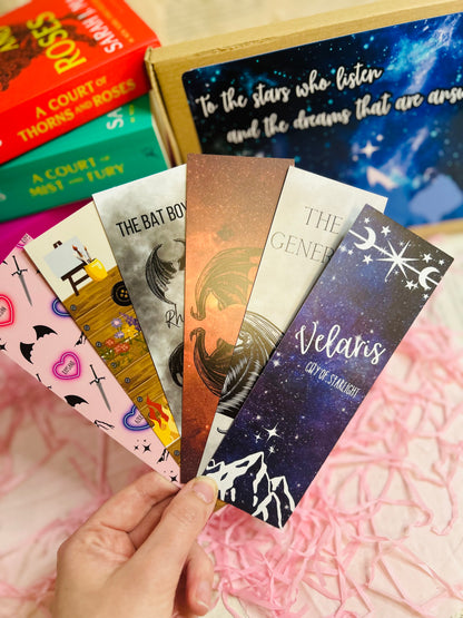 Flat lay of an ACOTAR-themed mystery bookish bundle featuring double-sided bookmarks, holographic stickers, a keyring, art print, annotate tabs, pen and highlighter. Items are displayed with Sarah J. Maas books and pink shredded paper, creating a cosy, fantasy-inspired aesthetic.