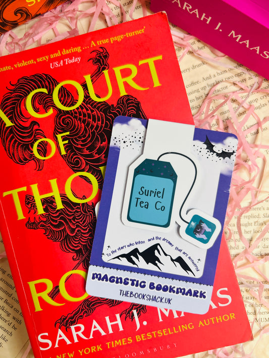 Blue tea bag-shaped magnetic bookmark labelled “Suriel Tea Co” clipped to an open fantasy novel, styled with pink shredded paper.