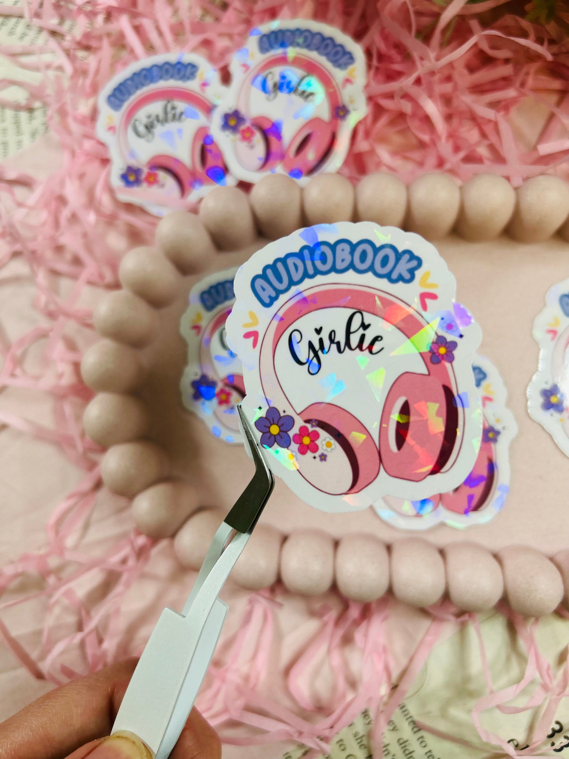 Holographic “Audiobook Girlie” sticker featuring pink headphones, colorful flowers, and sparkly finish — displayed with pastel decor and pink shredded paper.