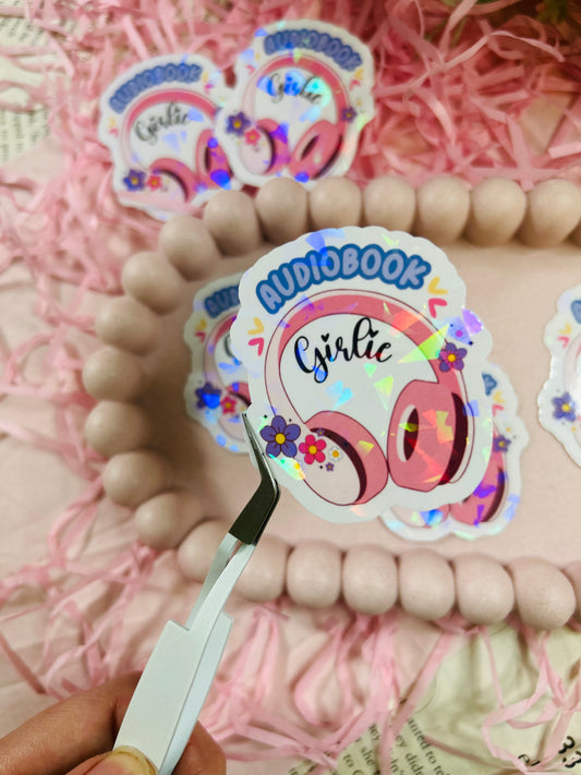 Holographic “Audiobook Girlie” sticker featuring pink headphones, colorful flowers, and sparkly finish — displayed with pastel decor and pink shredded paper.