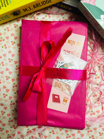  A pink bookish gift box wrapped with a red ribbon, featuring a bookmark that says "I run on coffee and spicy books." 