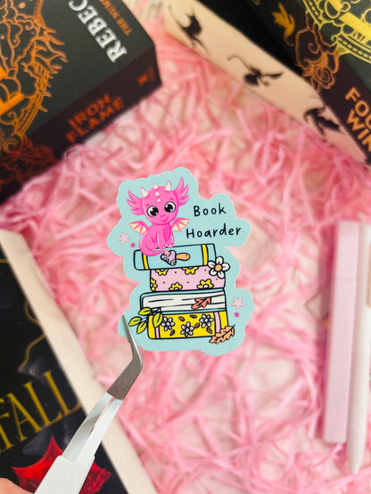 Close-up of a vibrant blue 'Book Hoarder' dragon sticker featuring a pink baby dragon perched on a stack of pastel-coloured books