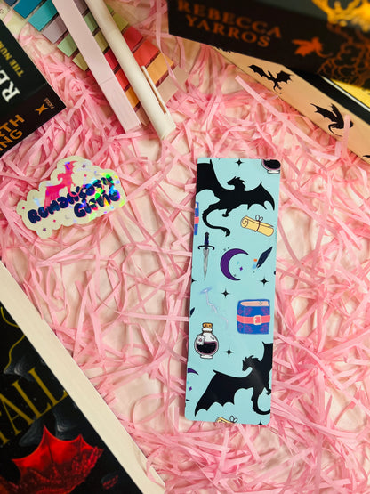 Fantasy-themed bookmark with a light blue background, showcasing a black dragon silhouette, potion bottle, sword, scroll, moon, and stars