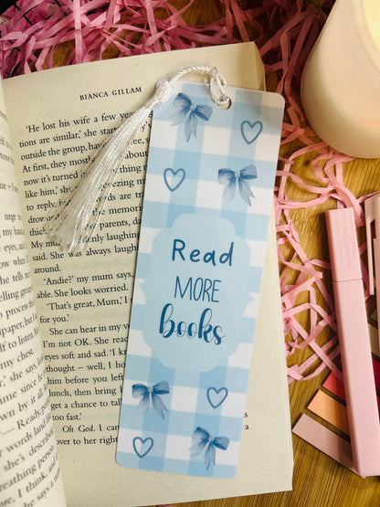 A blue gingham bookmark featuring elegant bow and heart designs, with the phrase "Read More Books" in blue lettering. The bookmark is accented with a soft white tassel and placed inside an open book, surrounded by pink shredded paper, a candle, and pastel stationery, creating a cosy bookish aesthetic.