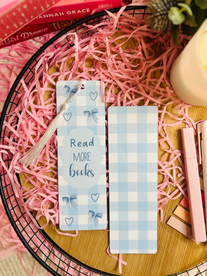 A blue gingham bookmark featuring elegant bow and heart designs, with the phrase "Read More Books" in blue lettering. The bookmark is accented with a soft white tassel and placed inside an open book, surrounded by pink shredded paper, a candle, and pastel stationery, creating a cosy bookish aesthetic.