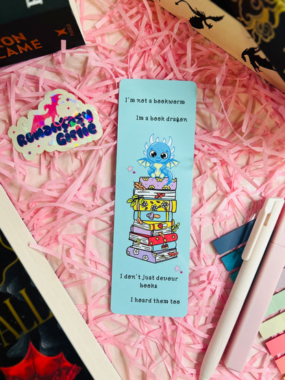 Whimsical blue bookmark featuring an adorable book dragon perched atop a colourful stack of books with the quote: 'I’m not a bookworm, I’m a book dragon. I don’t just devour books, I hoard them too.'