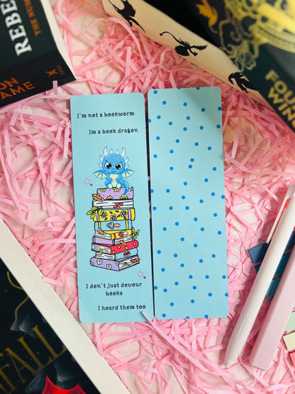 Charming two-sided blue bookmark set against pink shredded paper. One side features a whimsical design of a dragon sitting atop a vibrant stack of books, accompanied by the quote: 'I’m not a bookworm, I’m a book dragon. I don’t just devour books, I hoard them too.' The other side is decorated with playful blue polka dots, exuding a cheerful and light-hearted vibe.