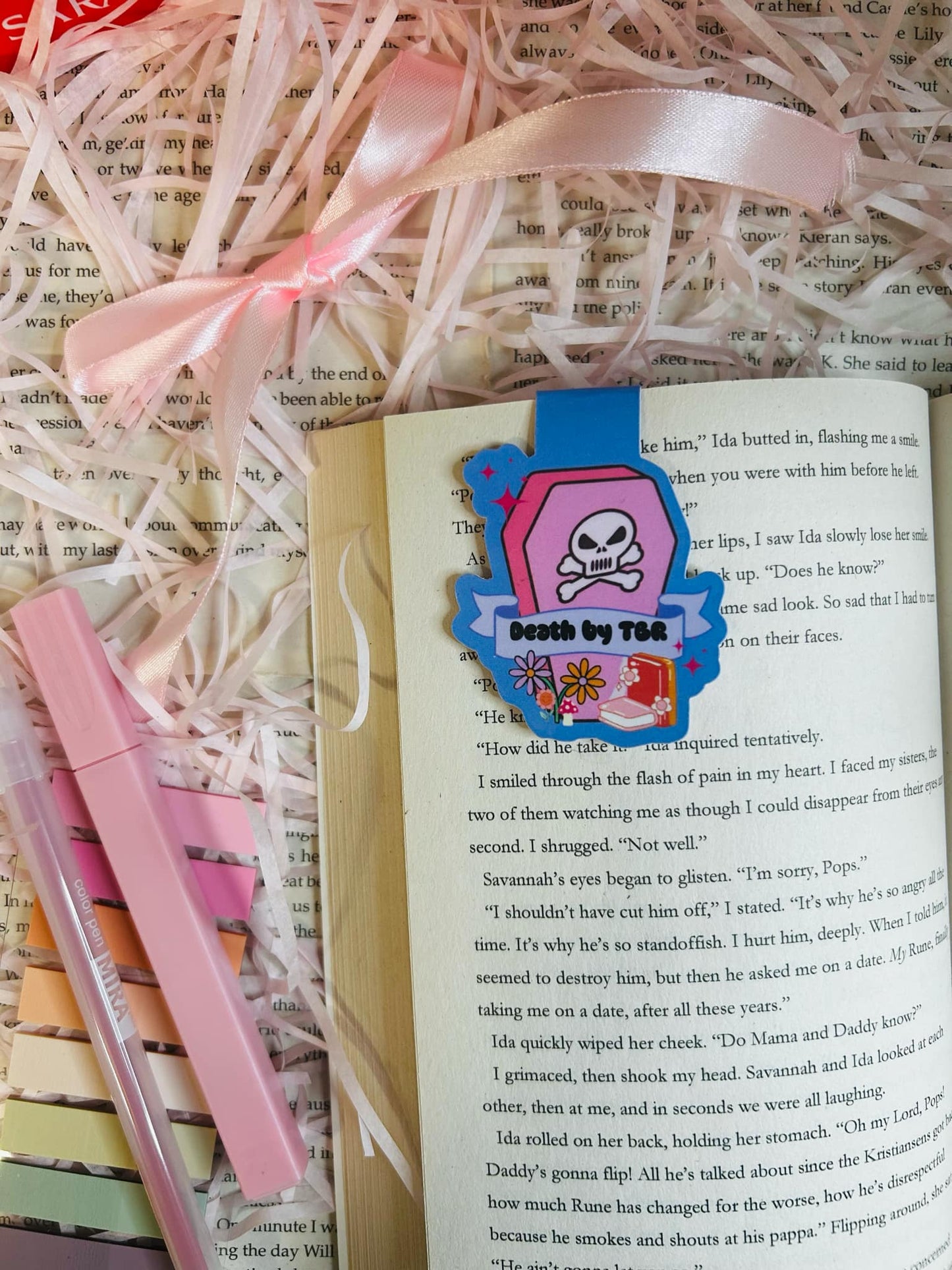 colourful 'Death by TBR' magnetic bookmark resting on an open book. The bookmark features a vibrant pink coffin design with a skull and crossbones, flowers, and a banner