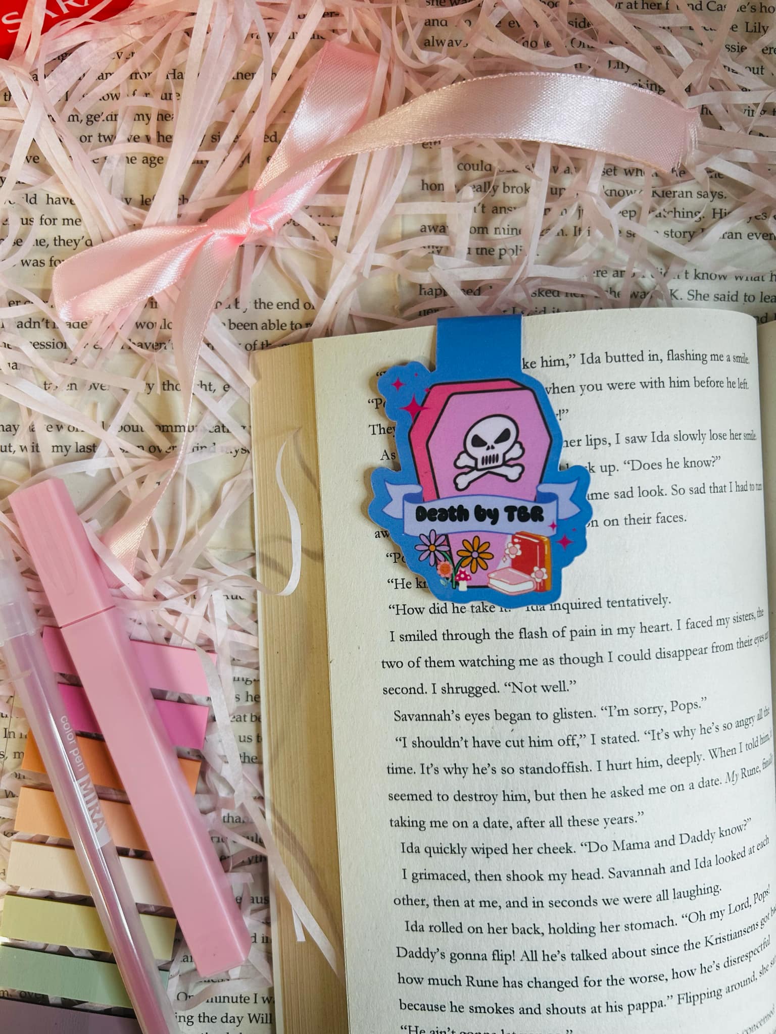 colourful 'Death by TBR' magnetic bookmark resting on an open book. The bookmark features a vibrant pink coffin design with a skull and crossbones, flowers, and a banner