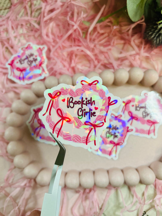 Holographic “Bookish Girlie” vinyl sticker with pink bows and rainbow shimmer, displayed with pink shredded paper and neutral wooden bead accents – perfect for aesthetic book lovers.