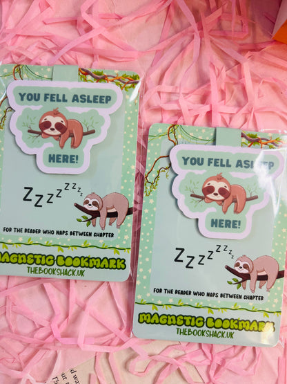 Sloth magnetic bookmark that says ‘You Fell Asleep Here’ placed on an open book with bookish stickers, pens, and novels in the background.