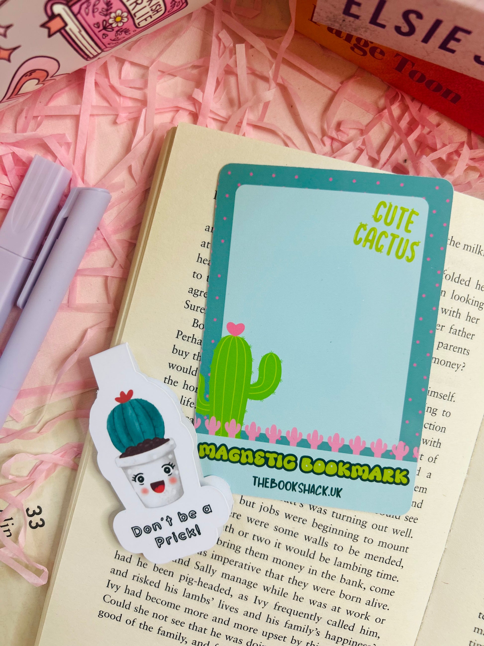 Funny cactus magnetic bookmark with the phrase ‘Don’t Be a Prick’ placed on an open book with pink shredded filler, pens, and books nearby.