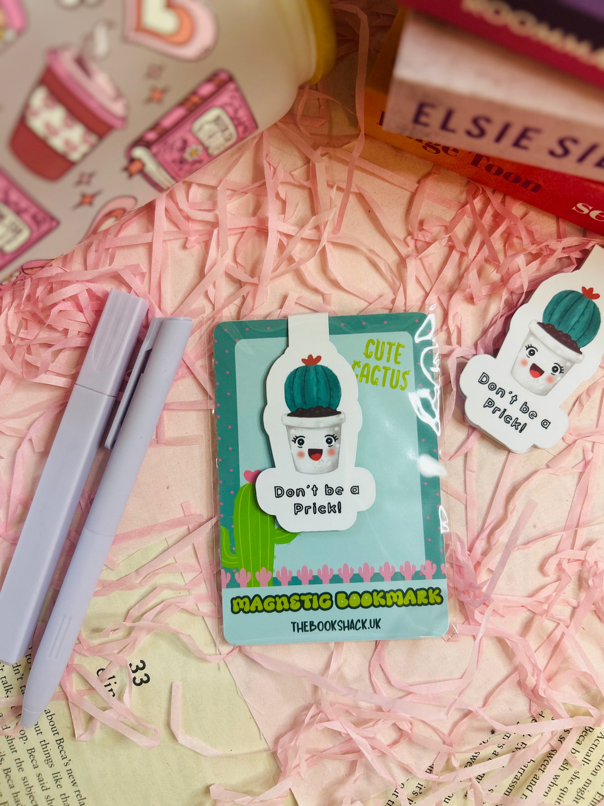 Funny cactus magnetic bookmark with the phrase ‘Don’t Be a Prick’ placed on an open book with pink shredded filler, pens, and books nearby.
