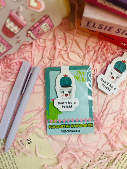 Funny cactus magnetic bookmark with the phrase ‘Don’t Be a Prick’ placed on an open book with pink shredded filler, pens, and books nearby.