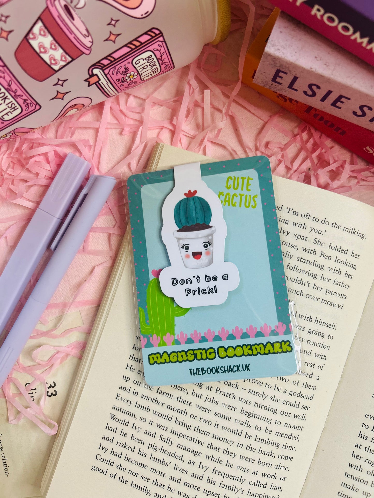 Funny cactus magnetic bookmark with the phrase ‘Don’t Be a Prick’ placed on an open book with pink shredded filler, pens, and books nearby.