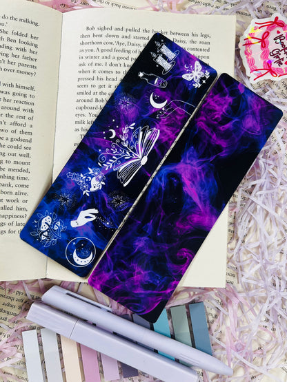 Dark purple witchy bookmark with celestial and magical illustrations including moons, candles, books, and stars, placed on an open book