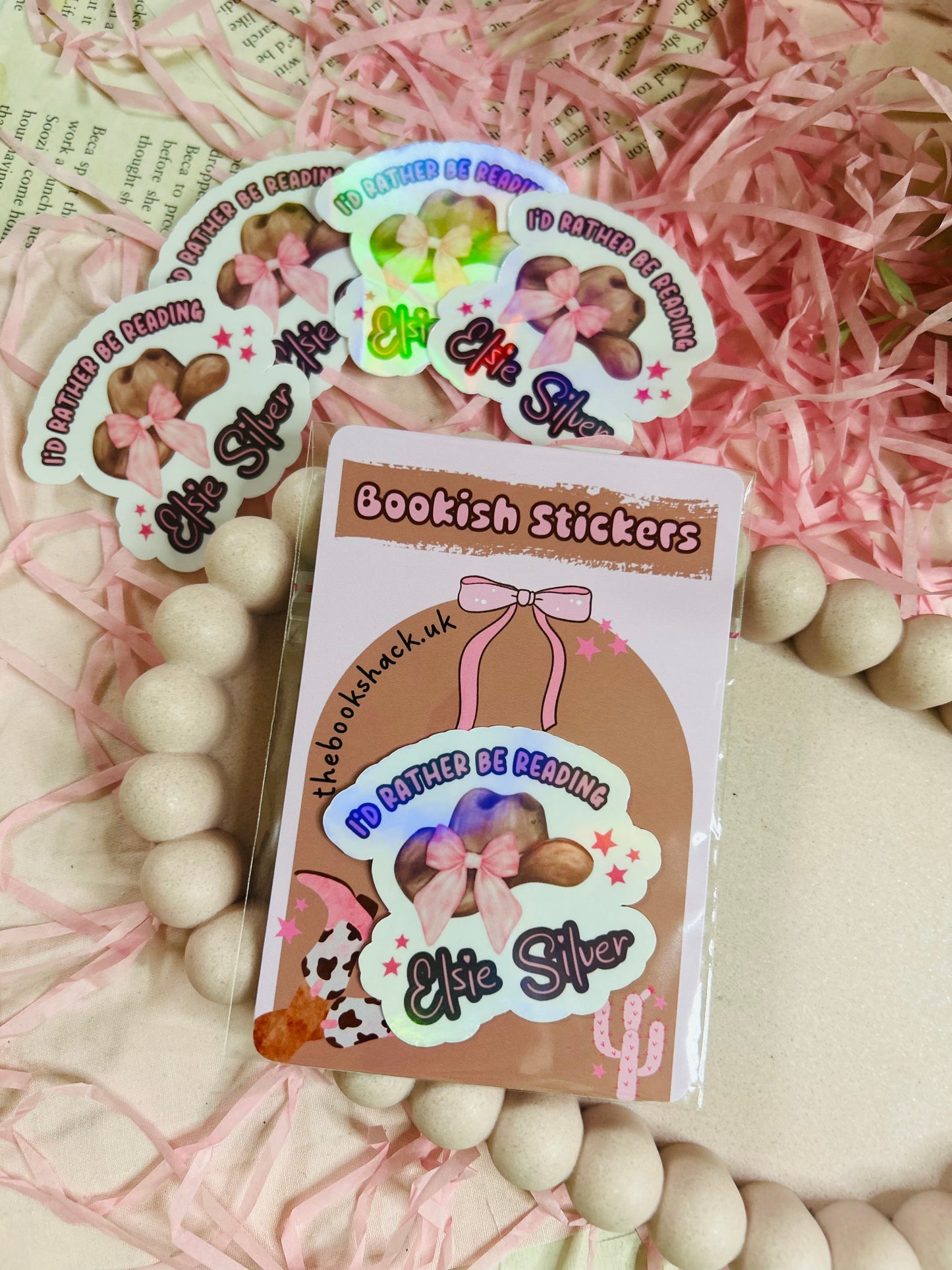 Holographic sticker with a pink-bowed cowboy hat and text reading "I'd Rather Be Reading Elsie Silver" on a bookish background with pink shredded paper