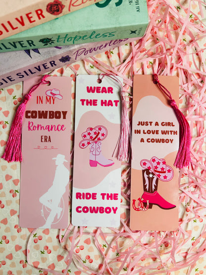 Set of three pink cowboy-themed bookmarks featuring tassels and playful designs, perfect for romance book lovers. Each bookmark showcases unique text like 'In My Cowboy Romance Era,' 'Wear the Hat, Ride the Cowboy,' and 'Just a Girl in Love with a Cowboy,' set against charming western-inspired illustrations of cowboy hats and boots. Ideal for marking pages in your favourite cowboy romance novels