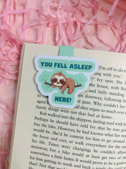 Sloth magnetic bookmark that says ‘You Fell Asleep Here’ placed on an open book with bookish stickers, pens, and novels in the background.