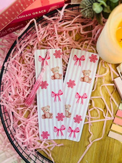 A pink teddy bear-themed bookmark with soft vertical stripes, delicate bows, and floral details, featuring a matching pink tassel. 