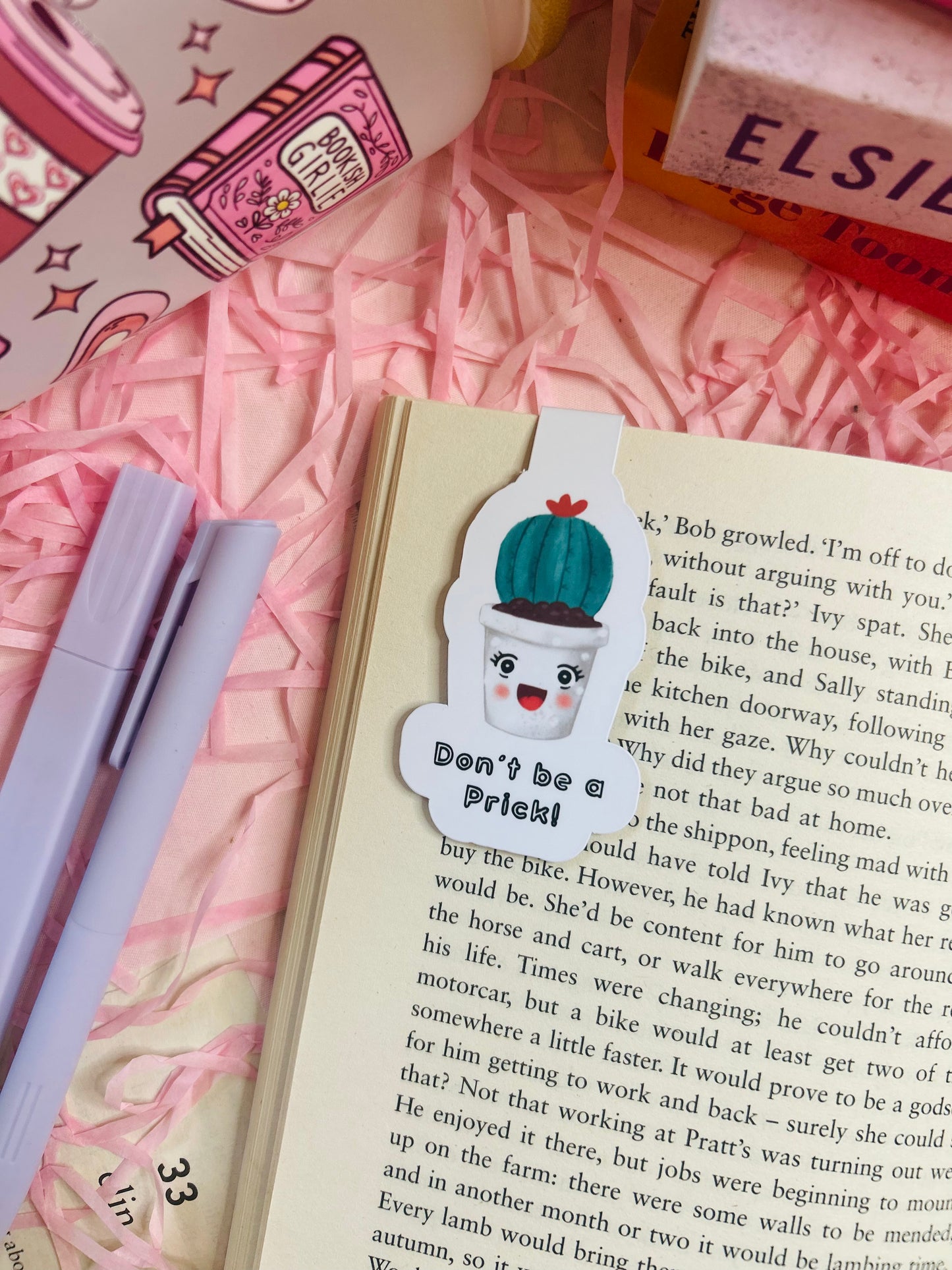 Funny cactus magnetic bookmark with the phrase ‘Don’t Be a Prick’ placed on an open book with pink shredded filler, pens, and books nearby.