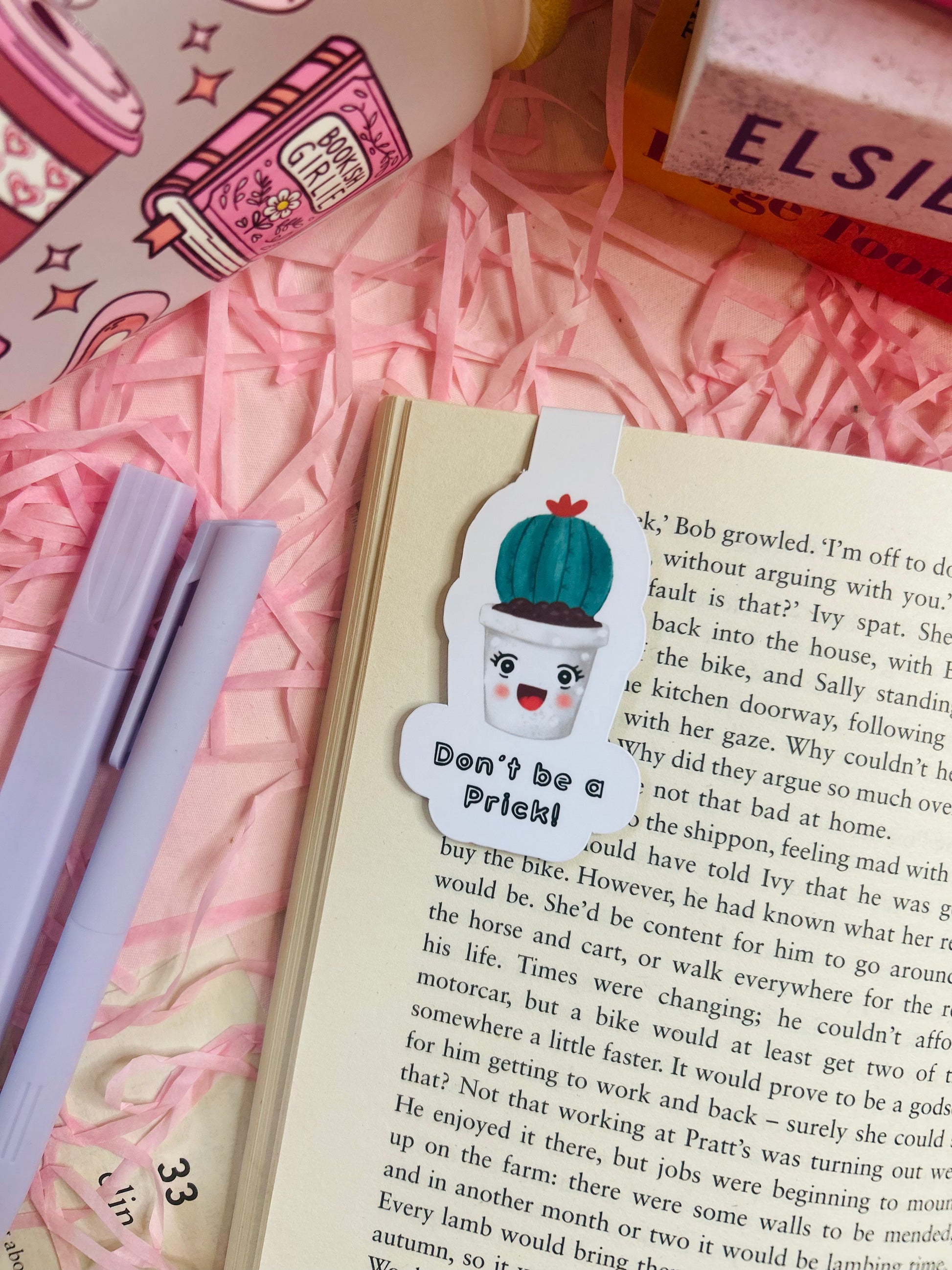 Funny cactus magnetic bookmark with the phrase ‘Don’t Be a Prick’ placed on an open book with pink shredded filler, pens, and books nearby.